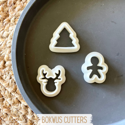Conjunto de 3 cortadores de massa em plástico branco com formas natalícias numa bandeja preta sobre tecido trançado