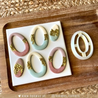 Moldes de cortadores ovais e de gota em plástico branco com detalhes coloridos e dourados, numa placa branca e bandeja de madeira.