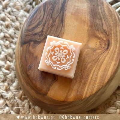 Molde de cortador quadrado com padrão floral branco sobre base de madeira oval e tecido de crochê