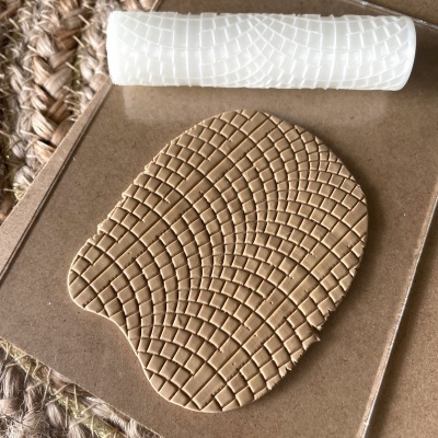 Rolo texturizador branco e massa bege com padrão quadriculado em ondas sobre cartão castanho