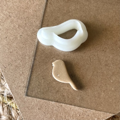 Cortador de madeira branco em forma de passarinho e peça recortada em madeira sobre cartão castanho