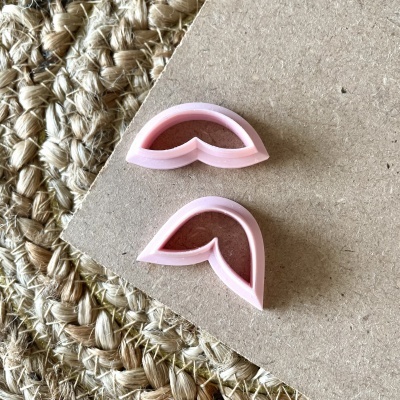 Dois cortadores de biscoitos rosa claro em forma de pétalas sobre cartão castanho e tapete de fibras naturais.