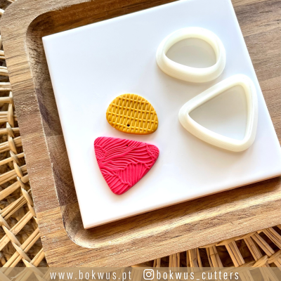 Cortadores triangulares brancos e massas texturizadas amarela e rosa sobre base branca e bandeja de madeira.