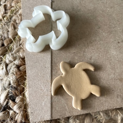 Cortador de biscoitos em plástico branco com biscoito cru em formato de tartaruga sobre cartão e tecido trançado.