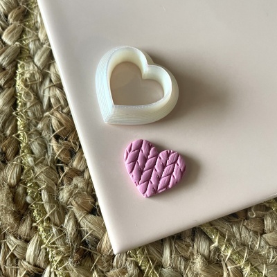 Cortador de biscoitos branco em forma de coração com biscoito rosa texturizado sobre superfície rosa claro e tapete de fibras naturais ao lado