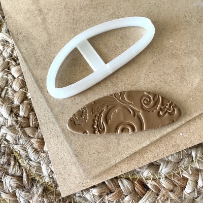 Cortador oval branco de plástico e pedaço de pasta castanha com relevo ornamental sobre superfície de madeira e tapete entrelaçado.