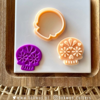 Três cortadores de massa em forma de caveira com padrões florais, em tons roxo e pêssego, sobre tabuleiro de madeira e placa branca.