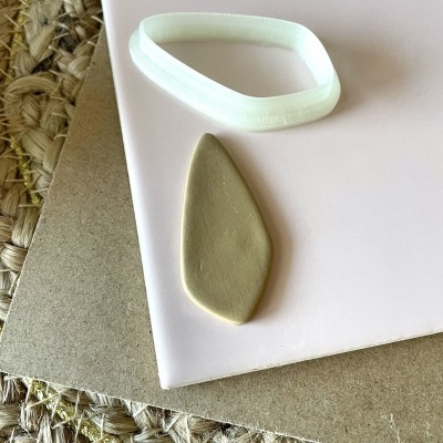 Pulseira elástica branca e peça de argila em forma de folha sobre superfícies claras e cartão com fundo de fibra natural.