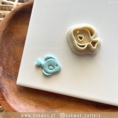 Dois cortadores de biscoitos em forma de peixe azul e bege sobre placa branca numa bandeja de madeira