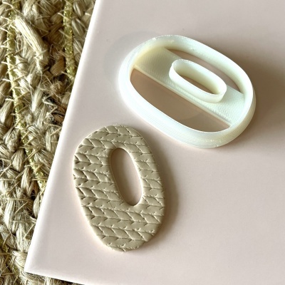 Dois objetos ovais, um branco em plástico e outro bege texturizado tipo pele, sobre superfície rosa clara junto a manta de corda entrelaçada.