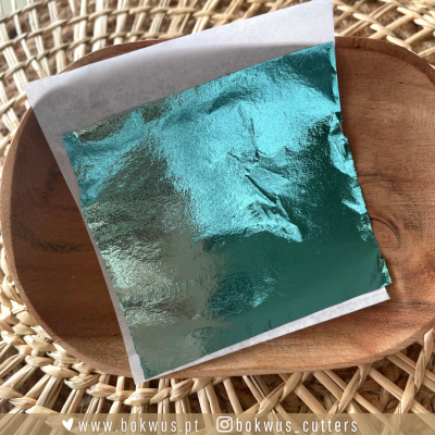 Lâmina metálica verde-azulada quadrada numa prato de madeira sobre base de vime.