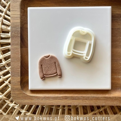 Corta-massas branco em forma de camisola ao lado de figura castanha em forma de camisola, sobre azulejo branco e bandeja de madeira