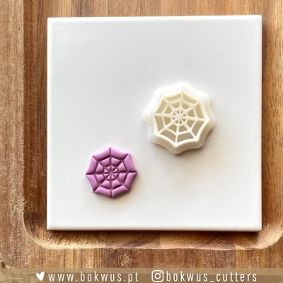 Dois cortadores de massa em forma de teia de aranha, um branco e outro rosa, sobre placa branca e madeira.