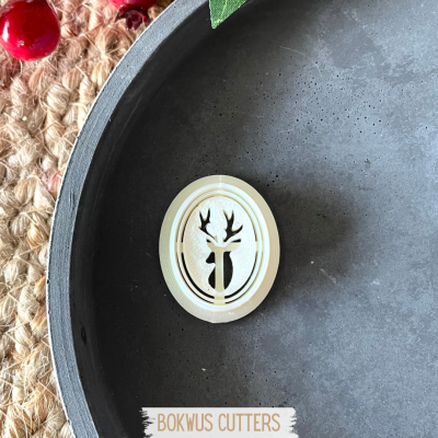 Cortador de bolachas oval branco com cabeça de veado recortada, sobre superfície escura, tecido rústico e frutos vermelhos no fundo