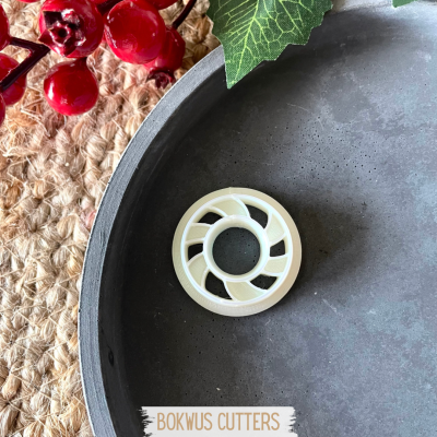 Cortador circular branco com design espiral dentro de recipiente metálico cinzento com decoração de plantas e tecido trançado