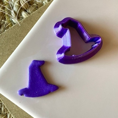 Cortador de bolachas roxo em forma de chapéu de bruxa com bolacha cortada da mesma forma e cor, sobre superfície branca com placa de cortiça e sisal