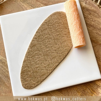 Rolo texturizador laranja com padrão ondulado sobre massa castanha com mesma textura em placa branca