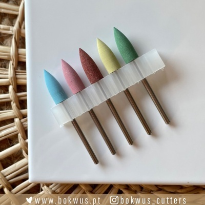 Conjunto de ponteiras de lixamento coloridas com hastes metálicas sobre caixa branca e fundo rattan