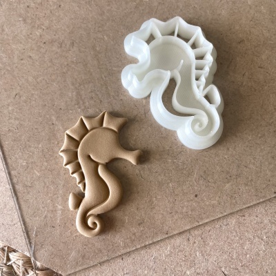 Dois cortadores de biscoitos em forma de cavalo-marinho, plástico branco e castanho, sobre superfície de madeira clara.