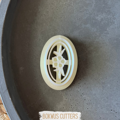 Cortante oval branco com padrão floral sobre superfície cinza escura.