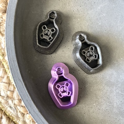 Três cortadores de biscoito em forma de garrafa com caveira, dois pretos e um roxo, numa tigela cinza