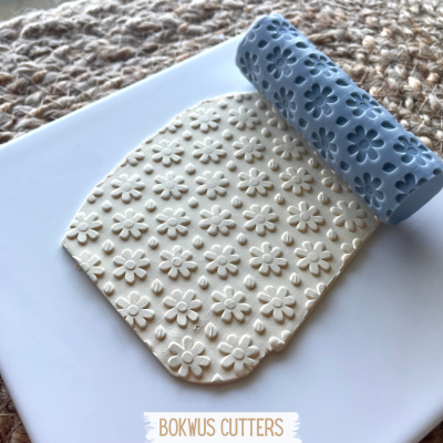 Rolo azul texturizado com padrão floral sobre massa branca impressa com o mesmo padrão, prato branco e tecido crochê ao fundo