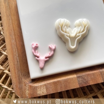 Dois moldes de corte em forma de cabeça de cervo, um rosa e um branco, sobre placa branca e tábua de madeira.