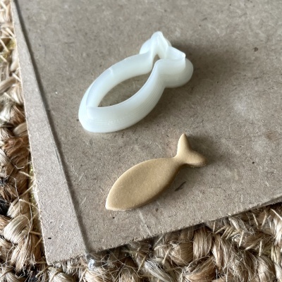 Cortador de biscoitos branco e biscoito dourado em forma de peixe sobre cartão castanho e tapete de fibras naturais