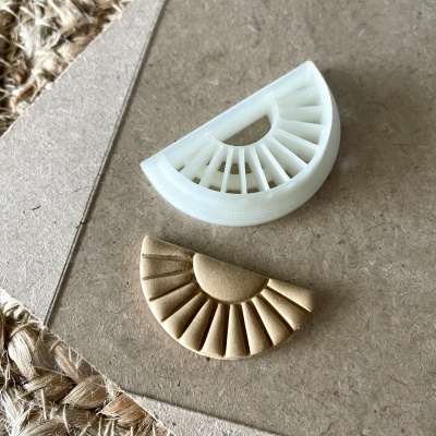 Dois objetos semicirculares, um branco translúcido e outro dourado com padrão em relevo, sobre superfície bege e tapete de fibras naturais.