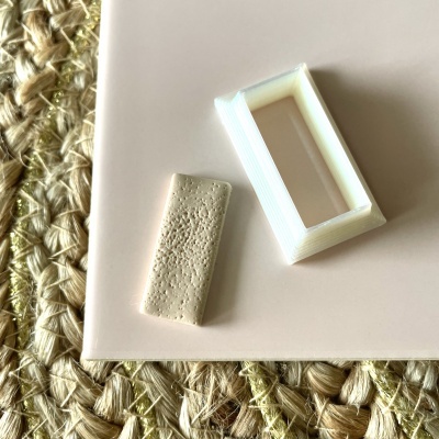 Duas peças retangulares, uma branca oca e outra bege texturada, sobre superfície bege e tapete de fibras naturais.