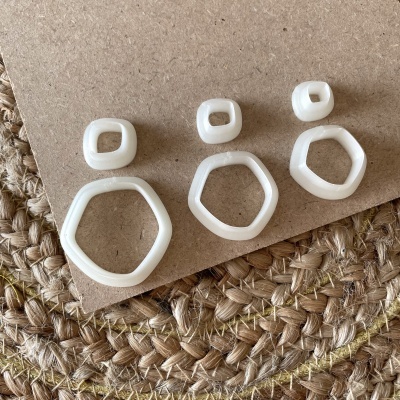 Três pares de brincos brancos geométricos sobre fundo castanho e tricot natural