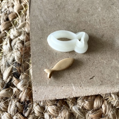 Dois objetos, um branco em forma de laço e outro bege em forma de peixe, sobre superfície castanha e tapete de juta.