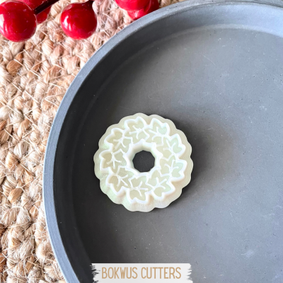 Cortante para bolachas branco com padrão de folhas sobre prato cinza e fundo de tecido trançado com bagas vermelhas.