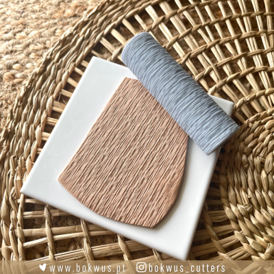 Rolo de fita cinza, cerâmica castanha texturizada e base branca sobre apoio de fibras naturais entrelaçadas