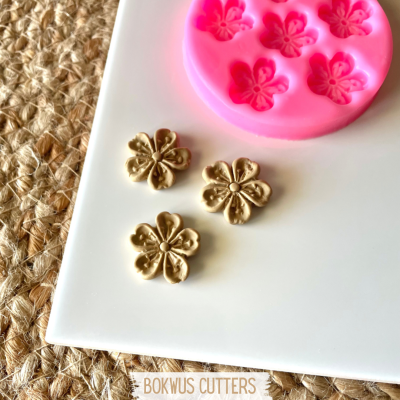 Cortantes de flores douradas e molde de silicone rosa para moldar flores sobre tabuleiro branco e pano de palha.