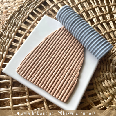 Massa de cerâmica bege e cinzenta com textura de linhas paralelas sobre placa branca e mesa de vime