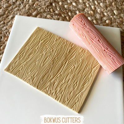 Rolo texturizador rosa com padrão de veios de madeira e placa retangular texturizada bege sobre superfície branca