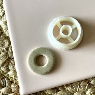 Duas peças circulares, uma cerâmica cinza e outra plástica branca, sobre superfície rosa e tapete trançado