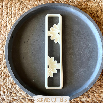 Cortador de pastelaria branco com design de flores amarelas, sobre bandeja cinzenta e tecido trançado