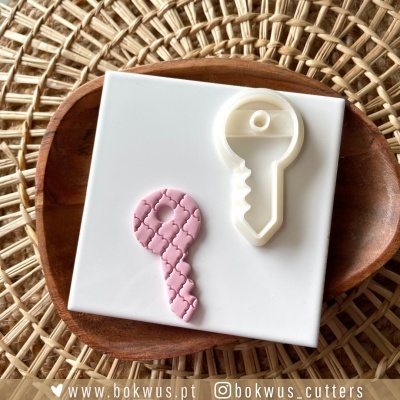 Cortador de massa em formato de chave branca com peça rosa texturizada em placa branca dentro de prato de madeira sobre vime.