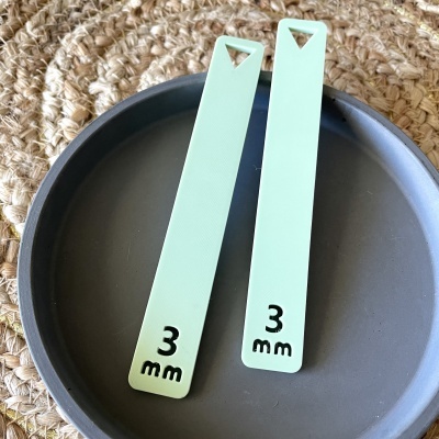 Dois marcadores de livro verdes claros com '3 mm' em preto, sobre superfície preta e base trançada.