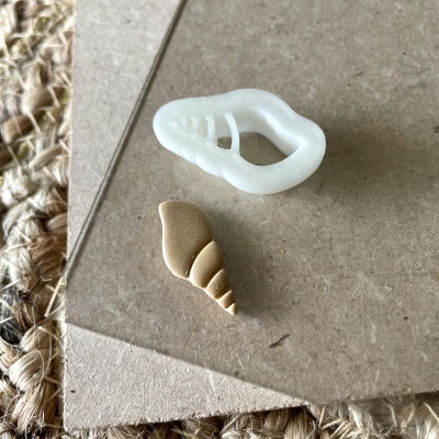 Duas peças pequenas, uma branca translúcida e outra bege com padrão de conchas, sobre superfície cinzenta e tapete de fibras naturais.