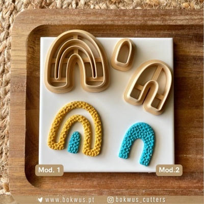 Cortadores de biscoitos em forma de arco-íris com texturas, dispostos em placa branca sobre fundo de madeira.