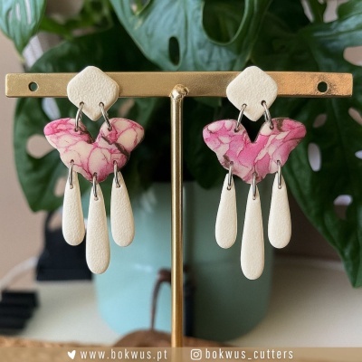 Brincos pendentes rosa e branco com padrão floral, em suporte dourado, planta verde ao fundo