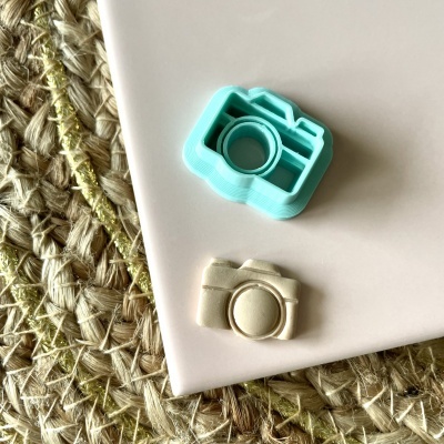 Dois moldes para biscoitos em formato de câmara fotográfica, azul turquesa e bege, sobre superfície branca e tapete de fibra natural.