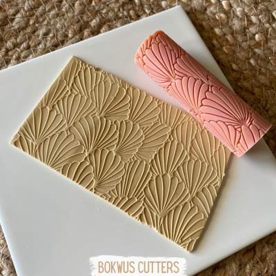 Rolo de textura rosa com padrão de conchas e placa bege estampada sobre base branca e fundo de tecido