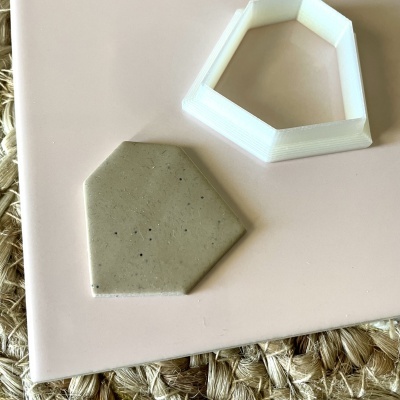 Molde pentagonal branco em plástico e peça pentagonal cinza em cerâmica sobre superfície rosa e tapete de fibras.