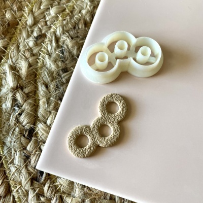 Dois objetos em forma de três círculos interligados, um branco e liso e outro bege e áspero, sobre superfície branca com fundo de tecido trançado.