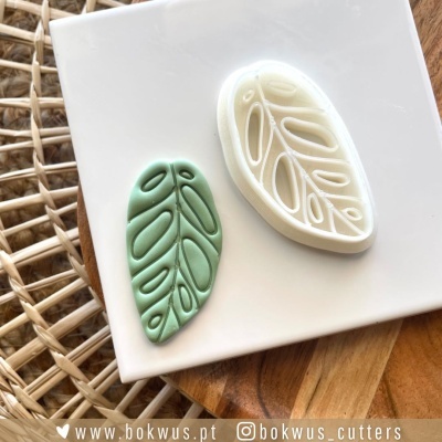 Dois cortadores de massa em forma de folha, um verde e outro branco, com padrões recortados, sobre base branca numa superfície de madeira e rattan.