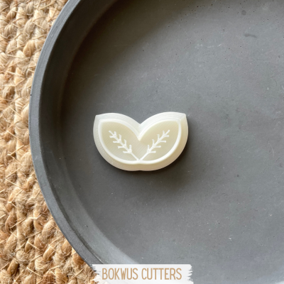 Cortador de bolachas branco em formato de folha com ramos brancos sobre prato cinzento e ráfia